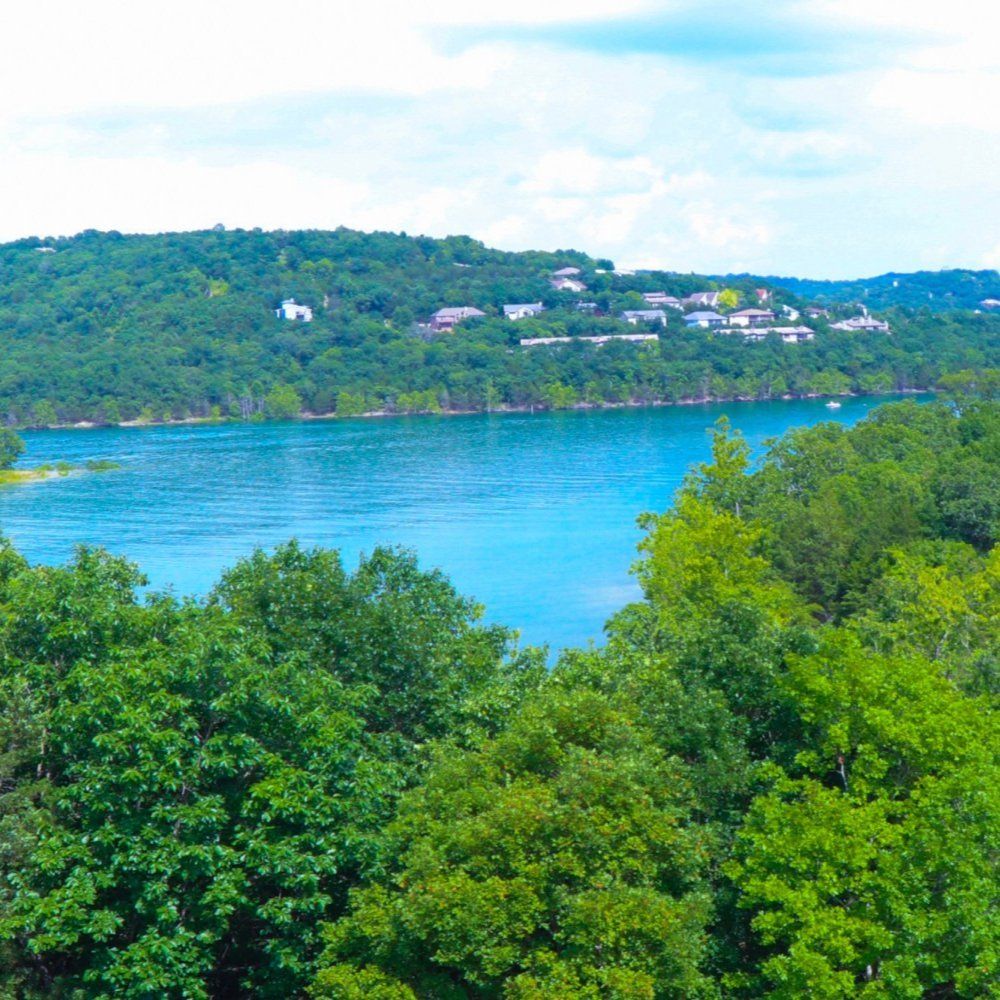 Lush green trees framing a blue lake with a forested hillside and scattered houses in the distance