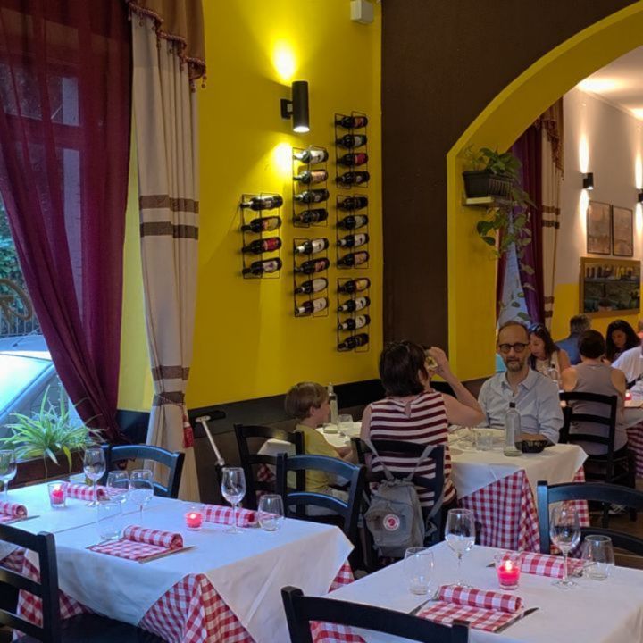 Interno di un ristorante con persone che cenano. Tavoli apparecchiati con tovaglie bianche e tovaglioli a quadretti rossi. Pareti gialle e marroni.