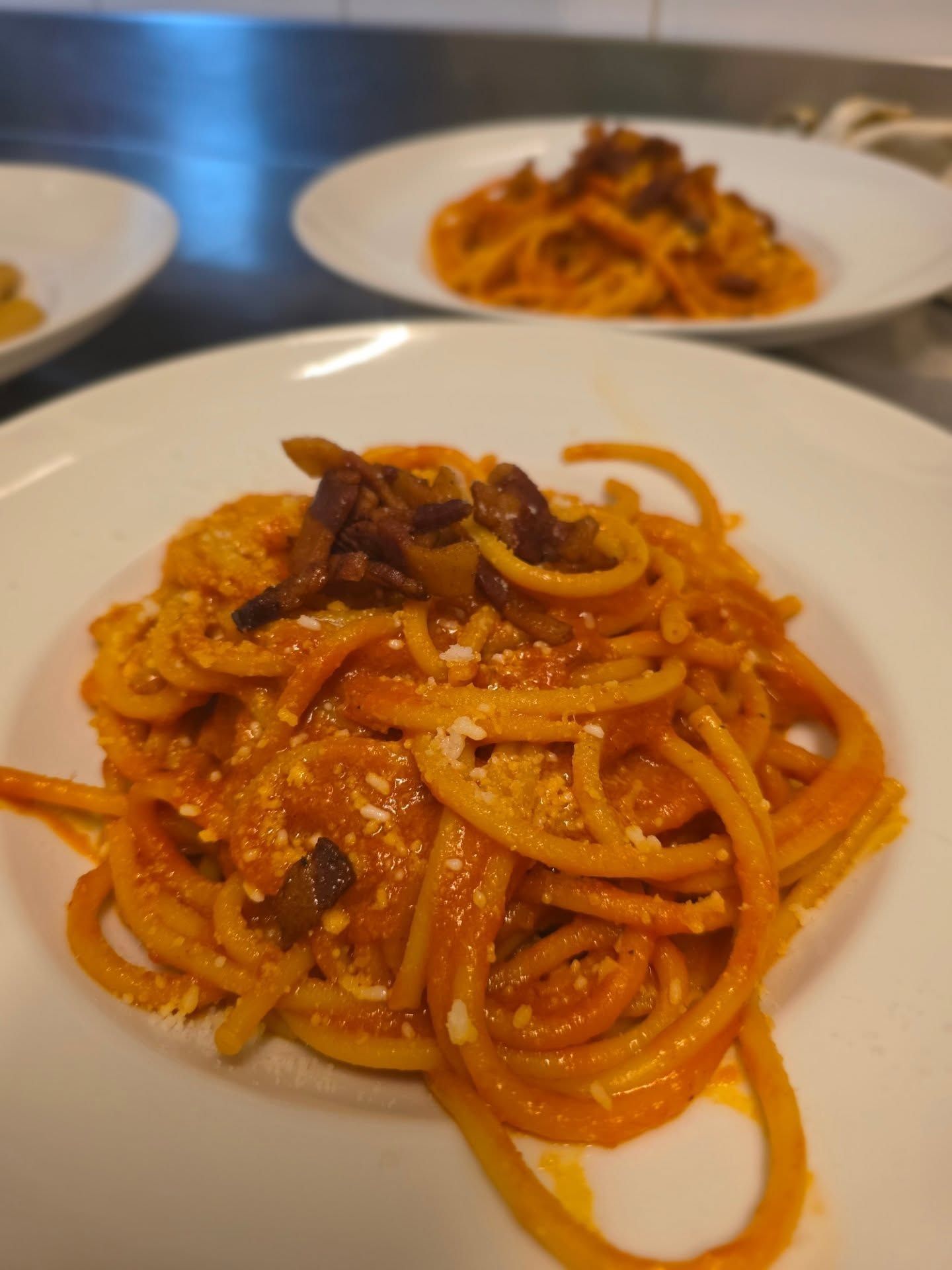 Piatto di spaghetti al sugo di pomodoro, guarnito con ingredienti diversi, servito su un piatto bianco; altri piatti sullo sfondo.