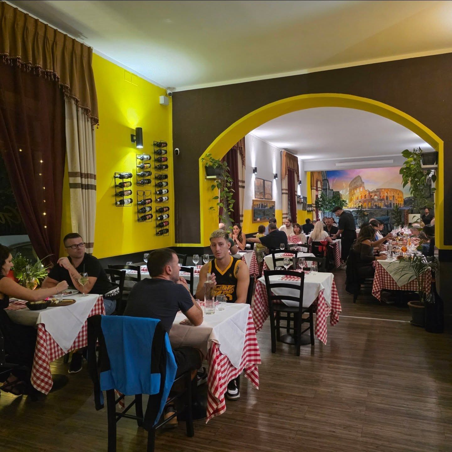 Interno del ristorante con commensali seduti ai tavoli, tovaglie a quadri rossi, pareti gialle e marroni, un ingresso ad arco.