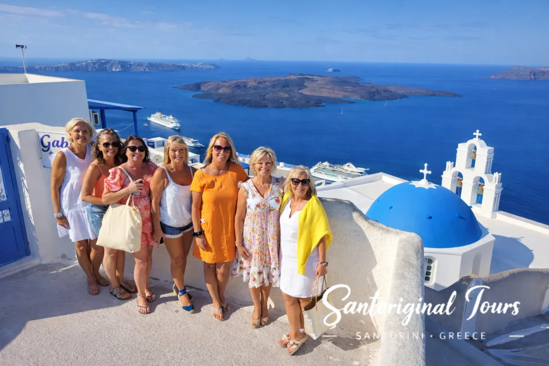 Group of women posing in Santorini with a blue dome church, caldera view, and cruise ships in the background