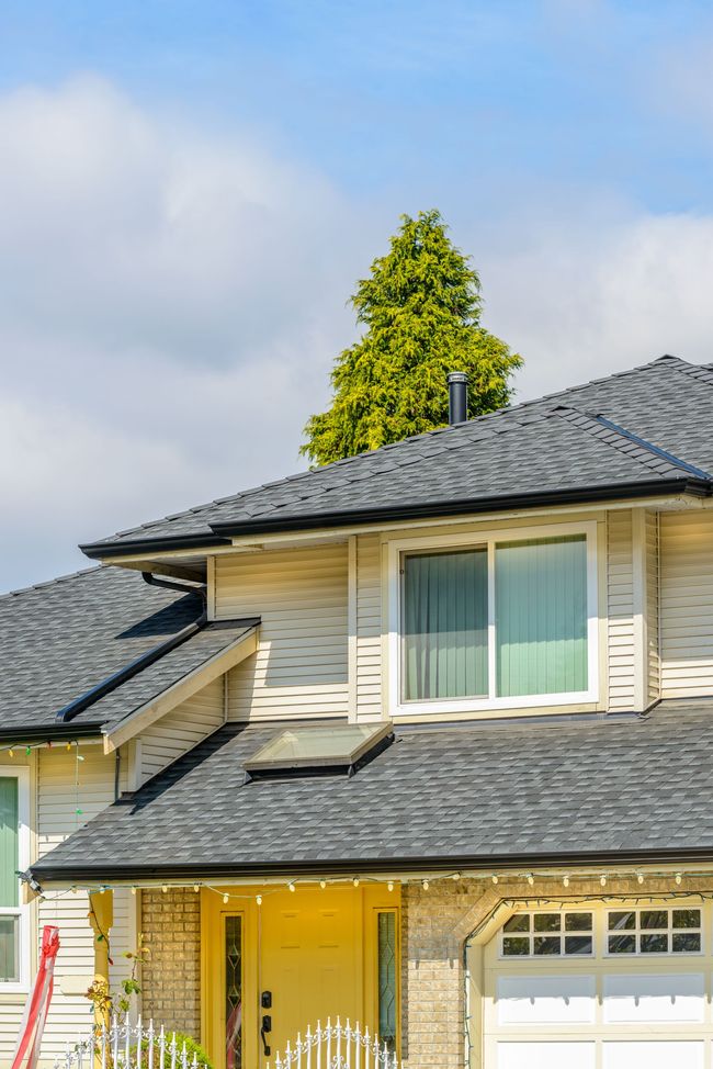 Two-story beige house with gray shingle roof and large front window under a blue sky