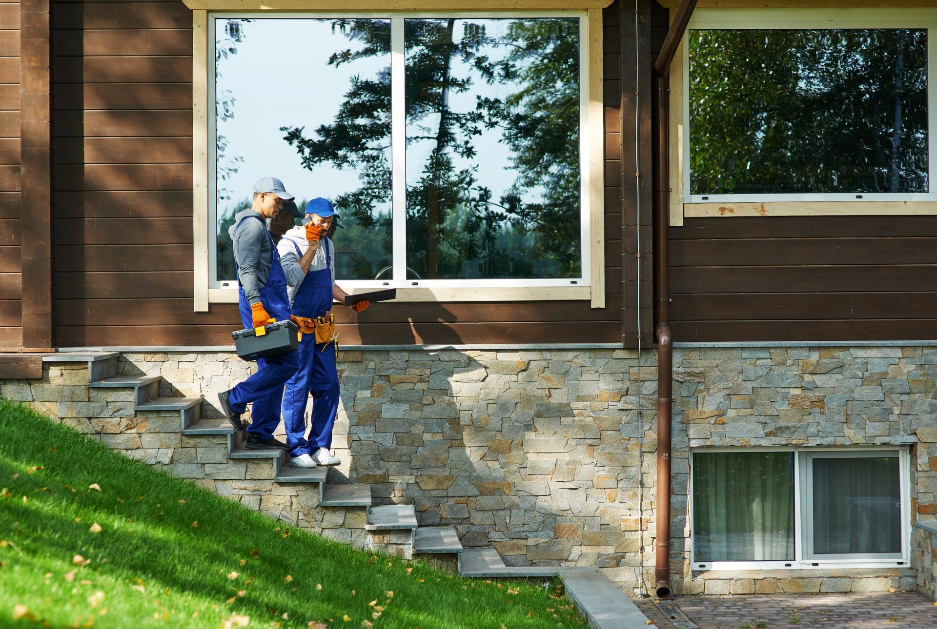 Two workers in blue uniforms carrying tools down stone steps beside a house.