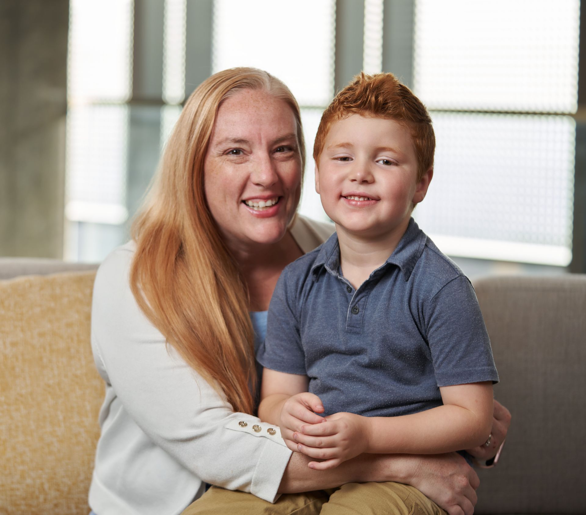 Charlie’s mother seated indoors holding Charlie on her lap, with soft natural light and windows in the background.