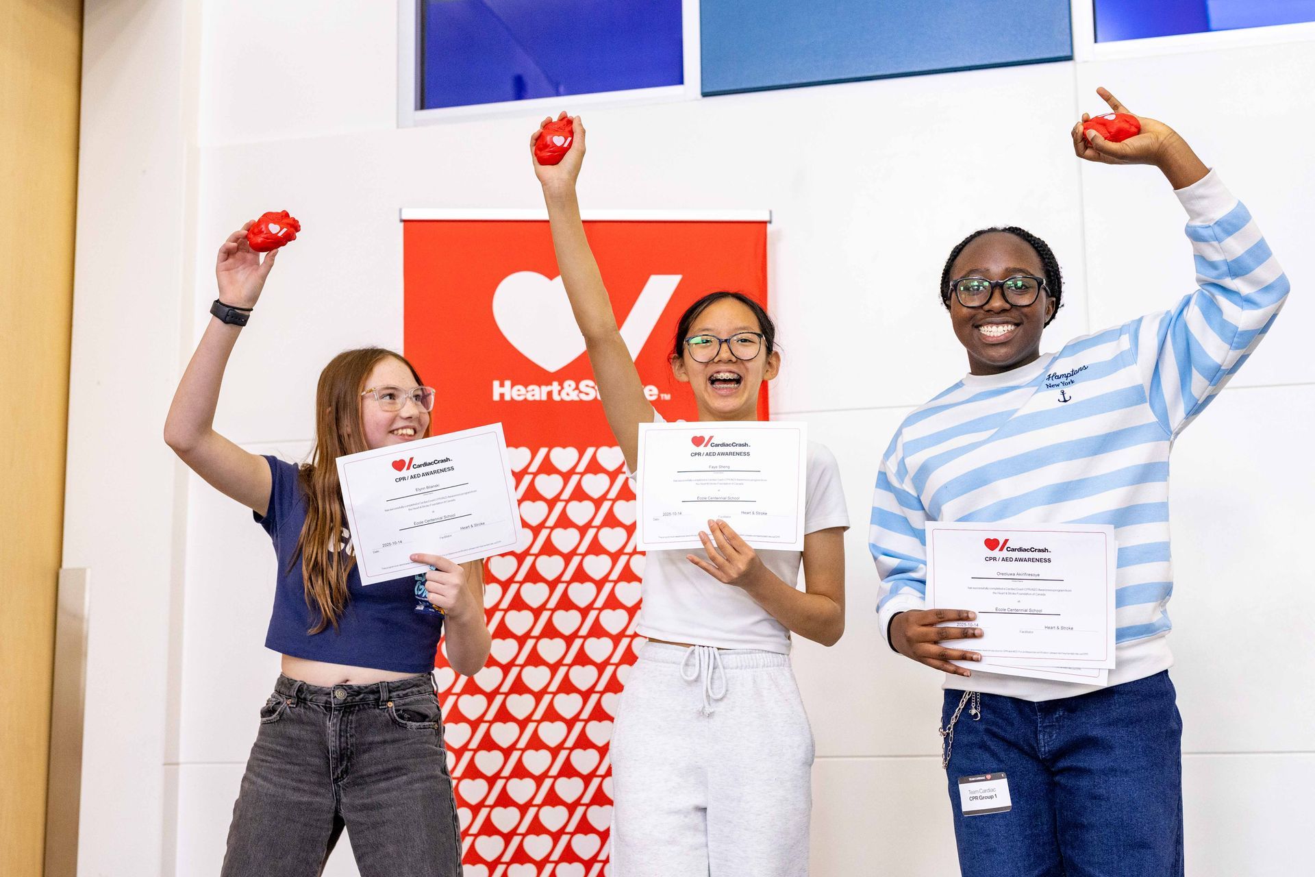 Students standing in front of a Heart & Stroke banner enthusiastically holding up their certificate of completion and heart shaped stress ball.