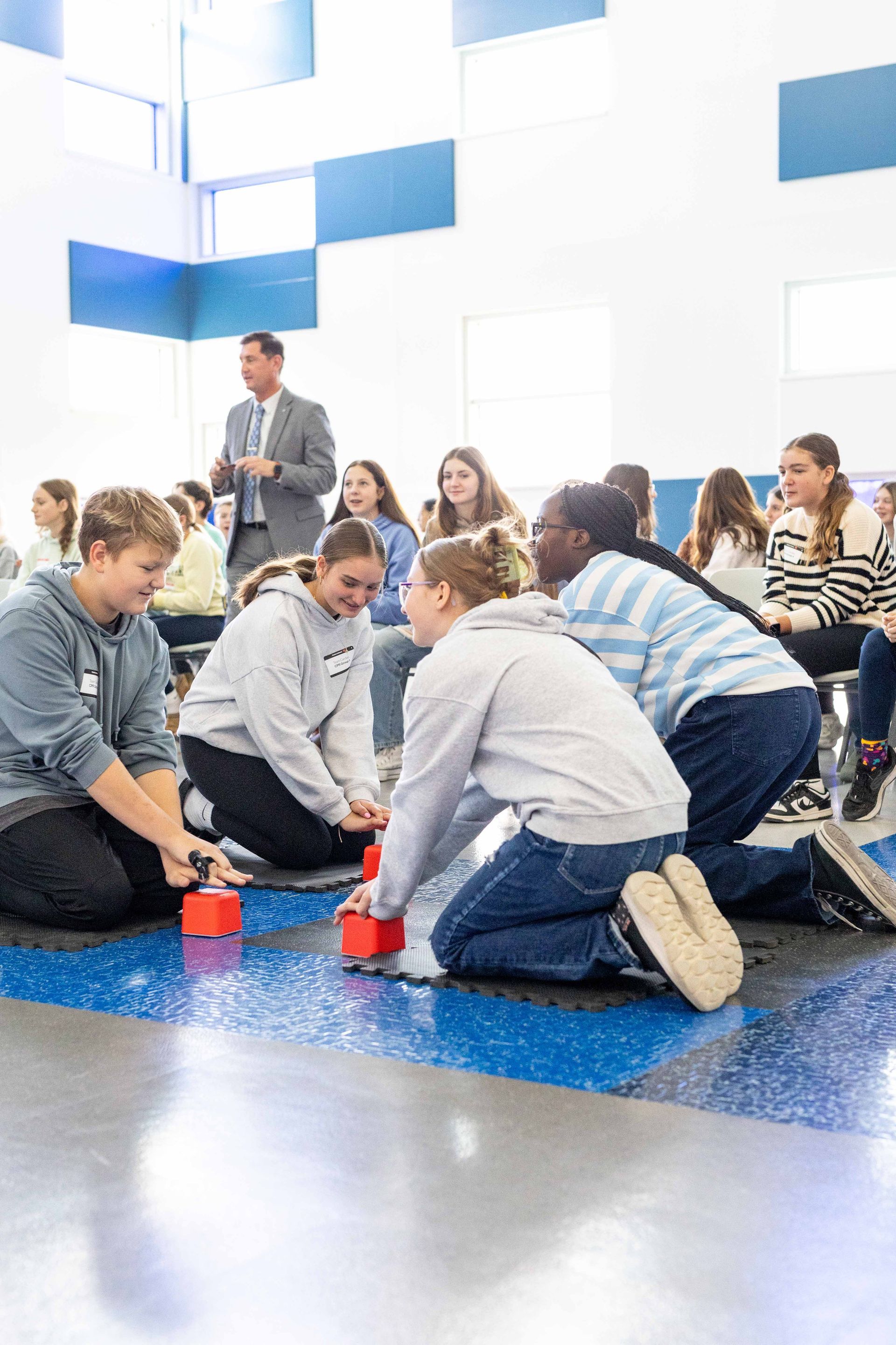 Students kneeling on the gymnasium floor with compression cubes practicing CPR chest compressions, while other students seated in chairs watch in the background.
