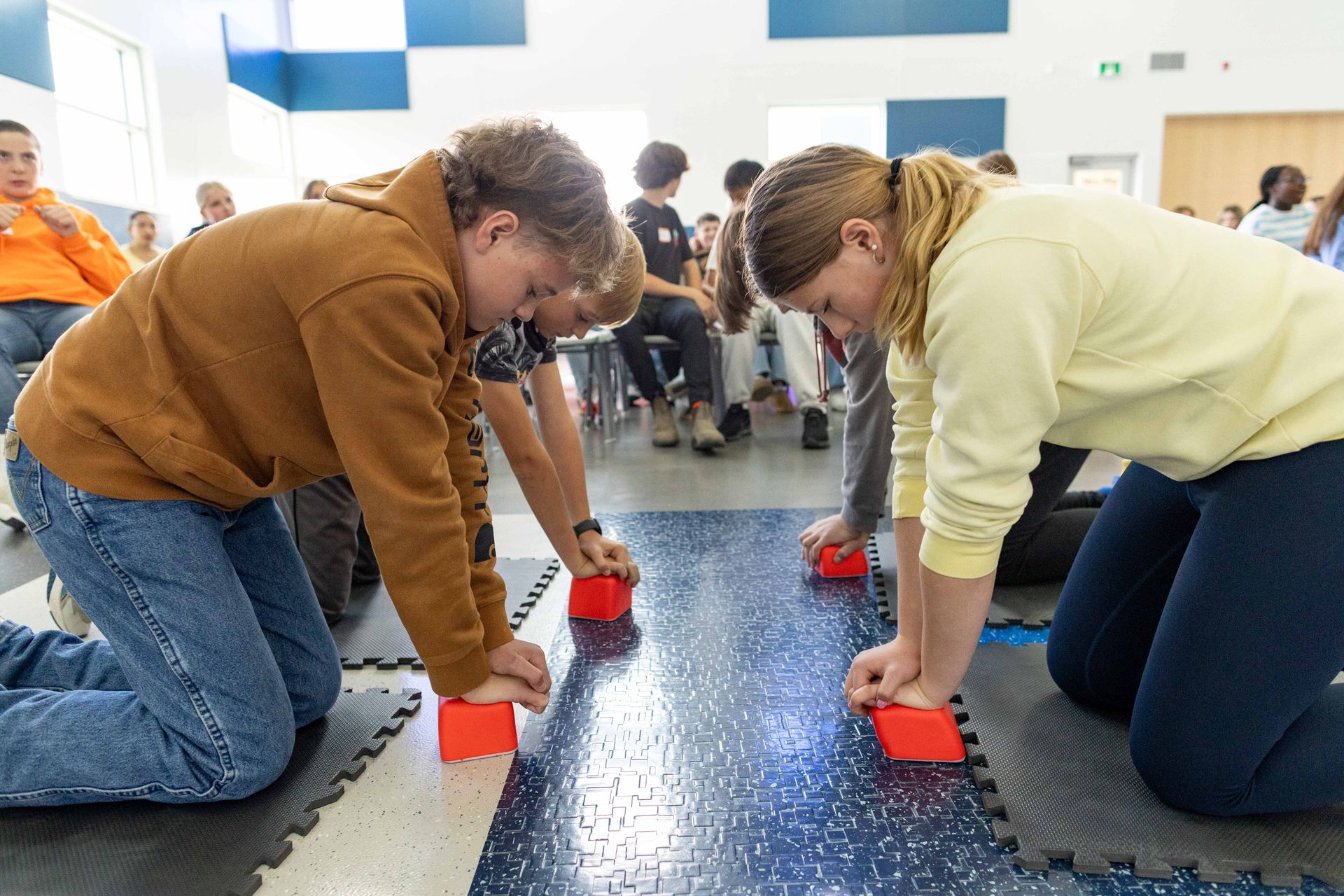 Students kneeling on the gymnasium floor with compression cubes practicing CPR chest compressions, while other students are seated watching the background.