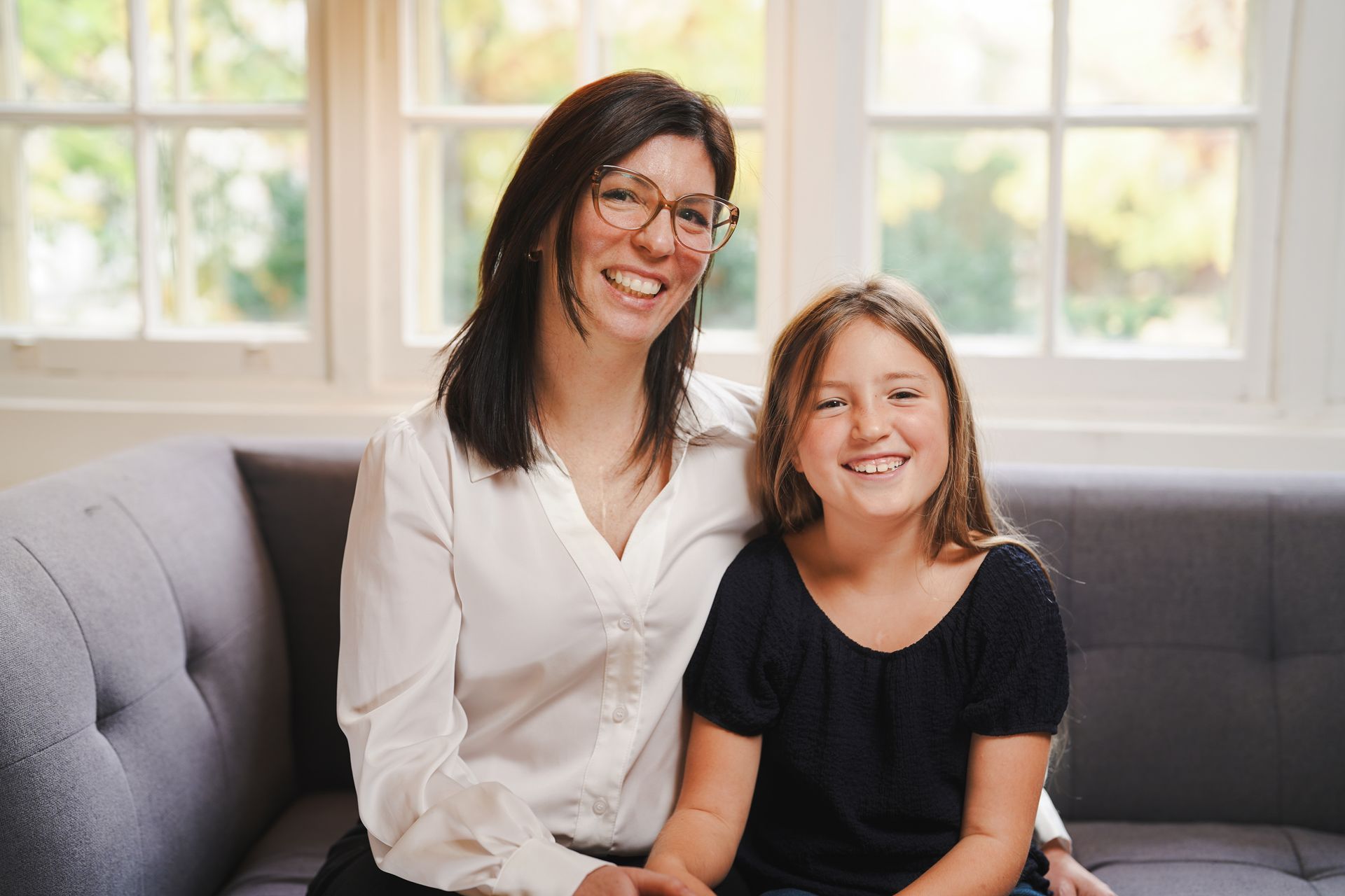 Sophie and Mia sitting side by side on a couch in a bright living room with large windows behind them.