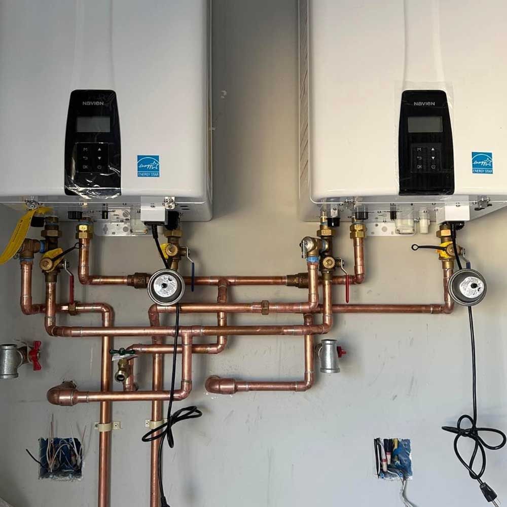 Two white tankless water heaters mounted on a wall with copper piping, valves, and gauges underneath.