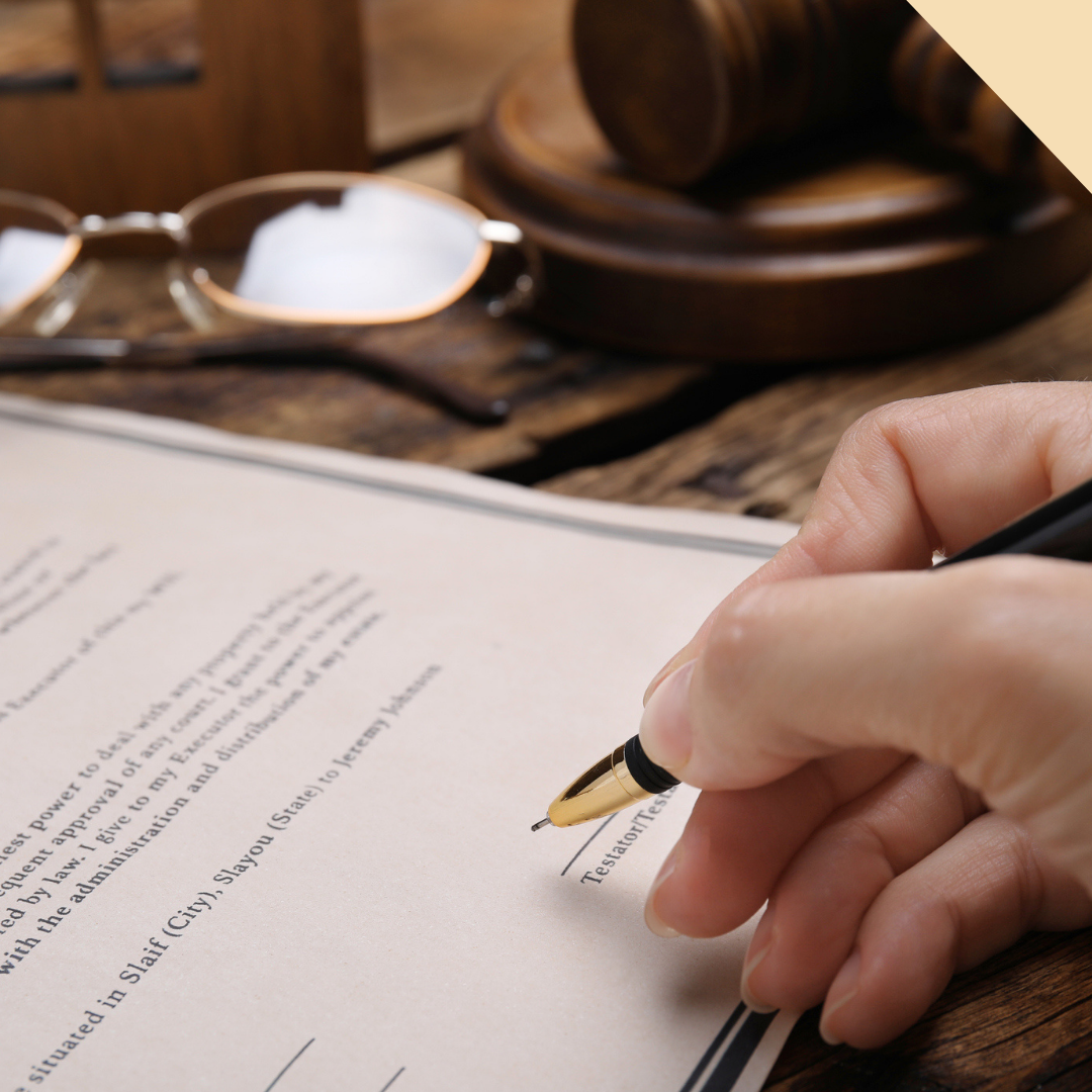 Hand signing a legal document with a pen, gavel, and glasses in the background.
