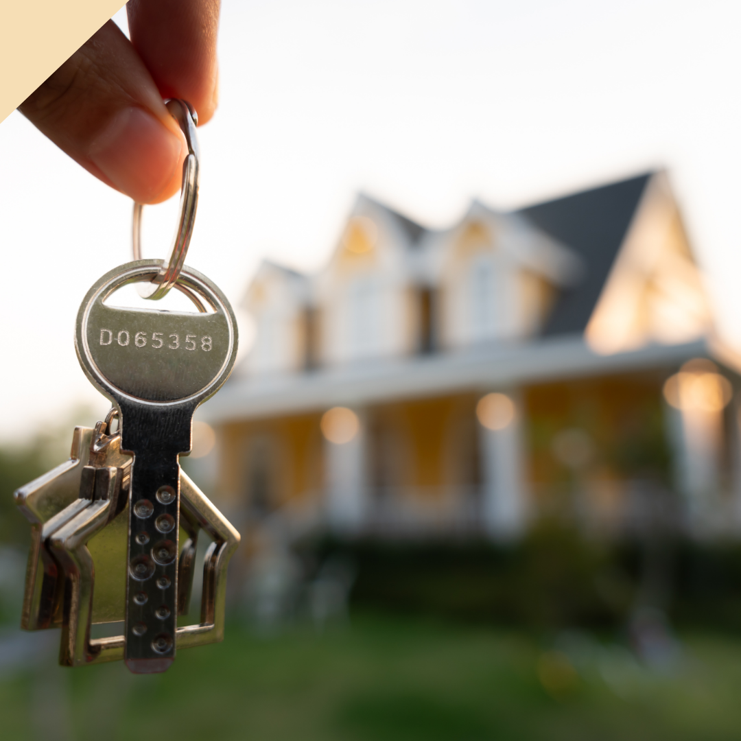 Hand holding keys with house-shaped fobs, blurred yellow house in background.