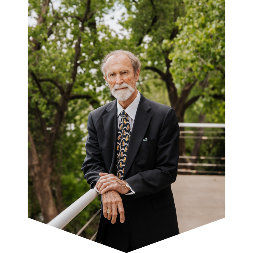 Man in a suit leans on a railing, smiling, trees in the background.