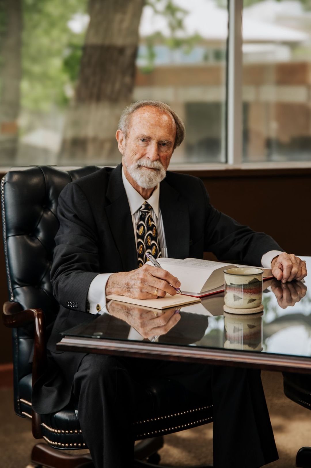An older person with a beard, wearing a suit and tie, sits at a desk writing in a book by a window.
