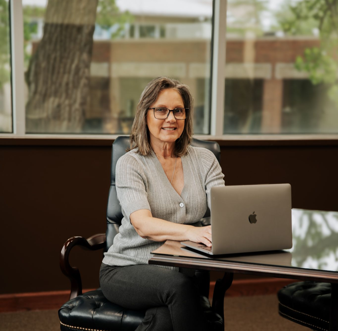 A person sits at a desk with a laptop in a bright office, looking toward the camera and smiling.
