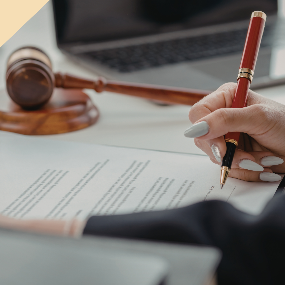 Person signing a document with a red pen, gavel and laptop on a desk.