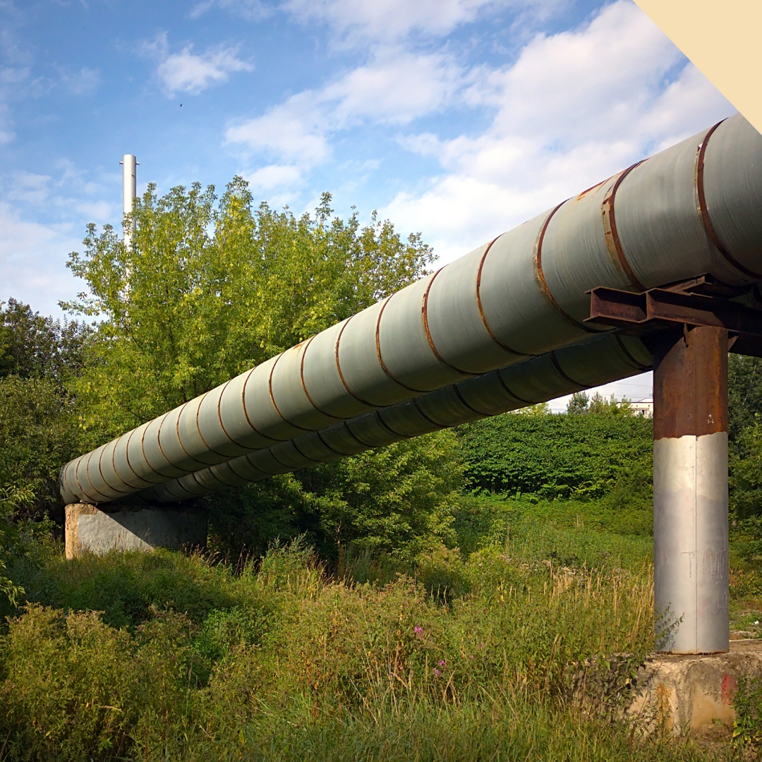Long, insulated industrial pipeline supported by metal posts, running through green foliage and under a blue sky.