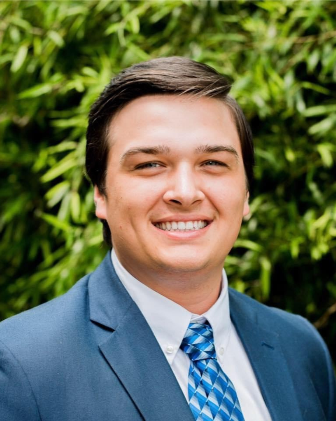 A smiling person in a blue suit and patterned blue tie standing in front of green foliage.