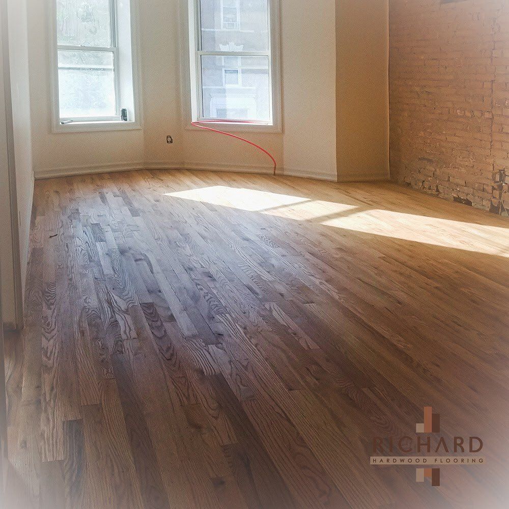 A bright room with newly installed hardwood flooring, two windows, and an exposed brick wall on the right side.