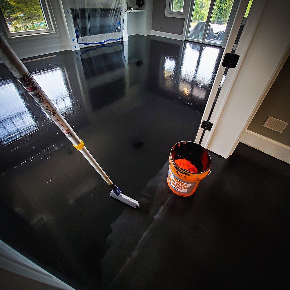 A high-gloss, black-painted floor in a room, with a paint roller and an orange bucket nearby.