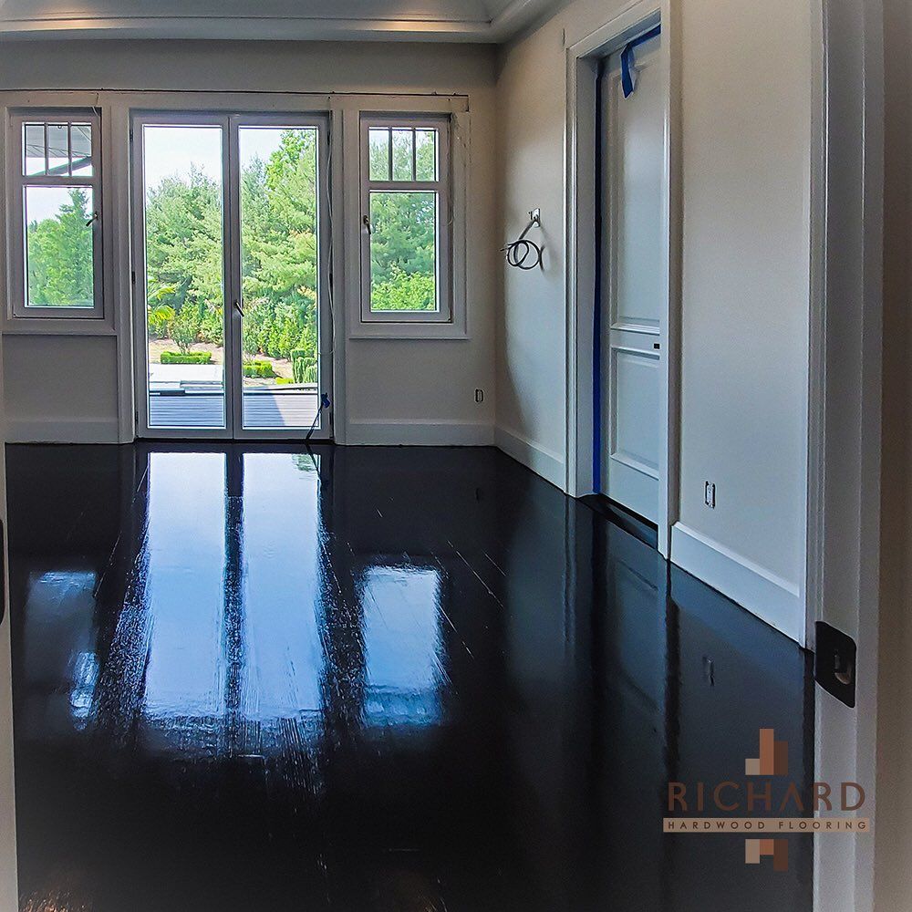 A room with high-gloss black hardwood floors, white walls, large windows, and French doors leading to a green yard.