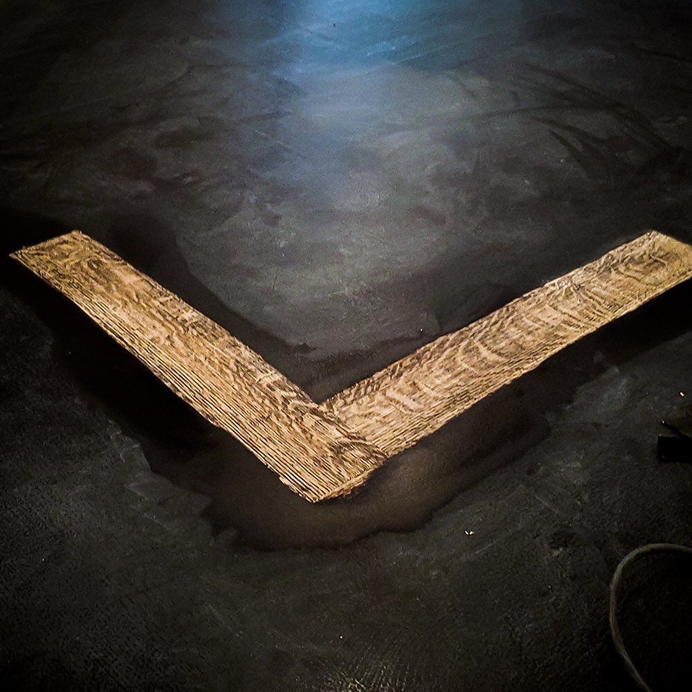 A V-shaped piece of light brown wood grain texture placed on a dark, textured floor.