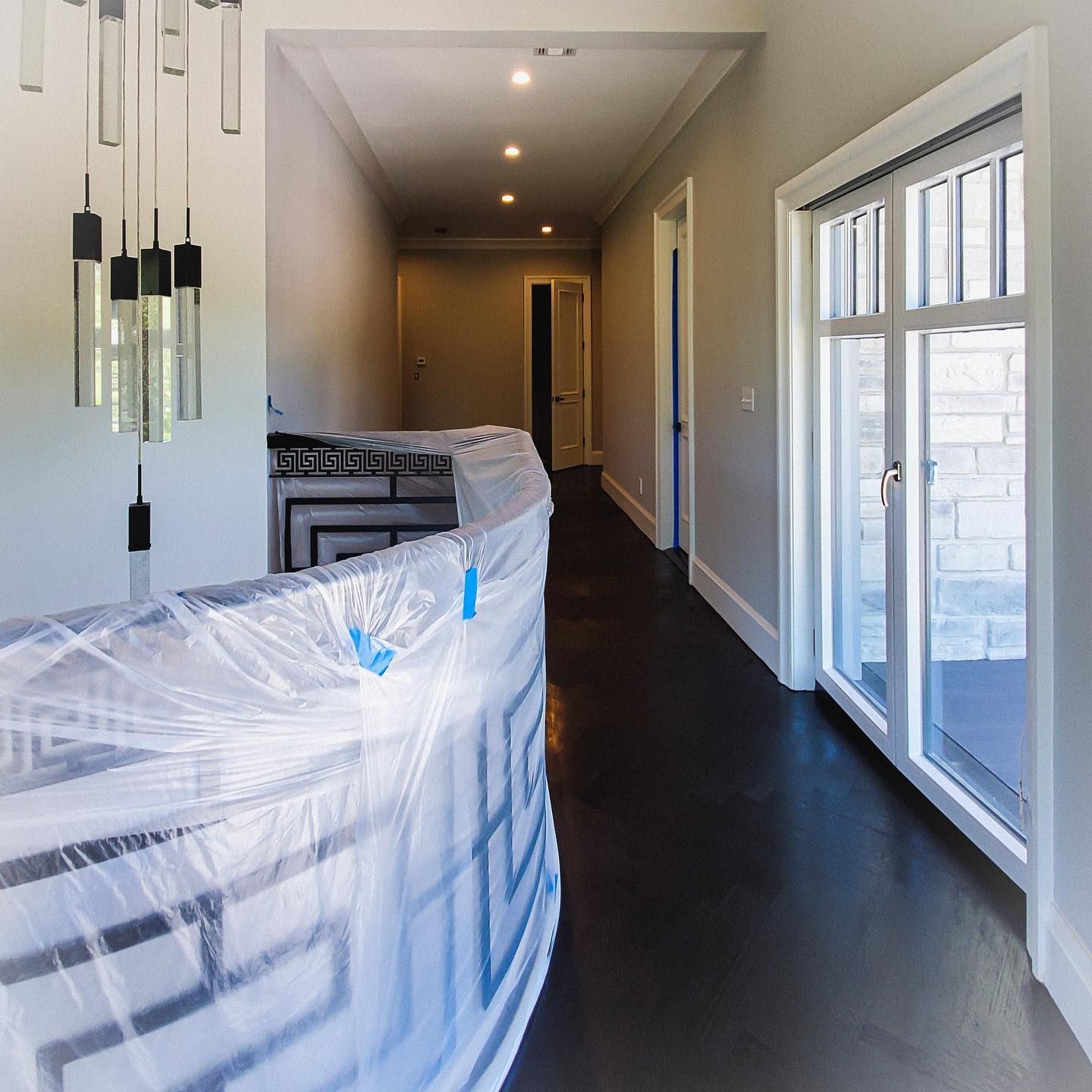 A hallway with dark hardwood floors, a staircase covered in protective plastic, and glass doors opening to the outdoors.