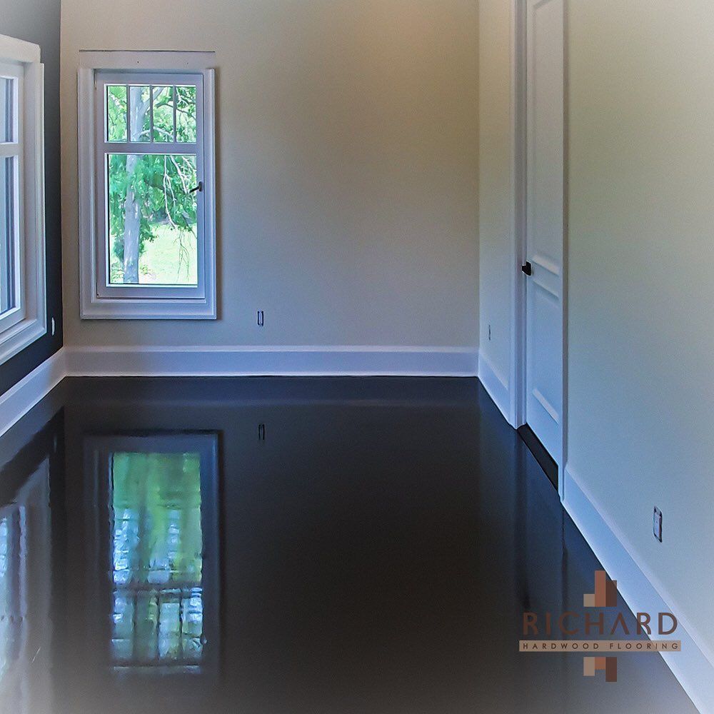 A bright, minimalist room featuring high-gloss, reflective black flooring, light-colored walls, and a window with white trim.