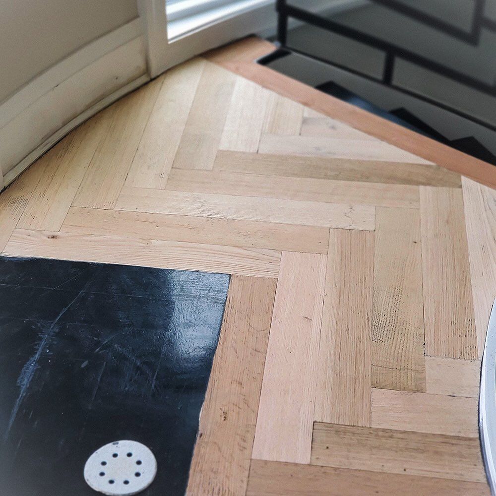 Light-colored wood flooring in a herringbone pattern next to a black floor area with a white circular drain cover.