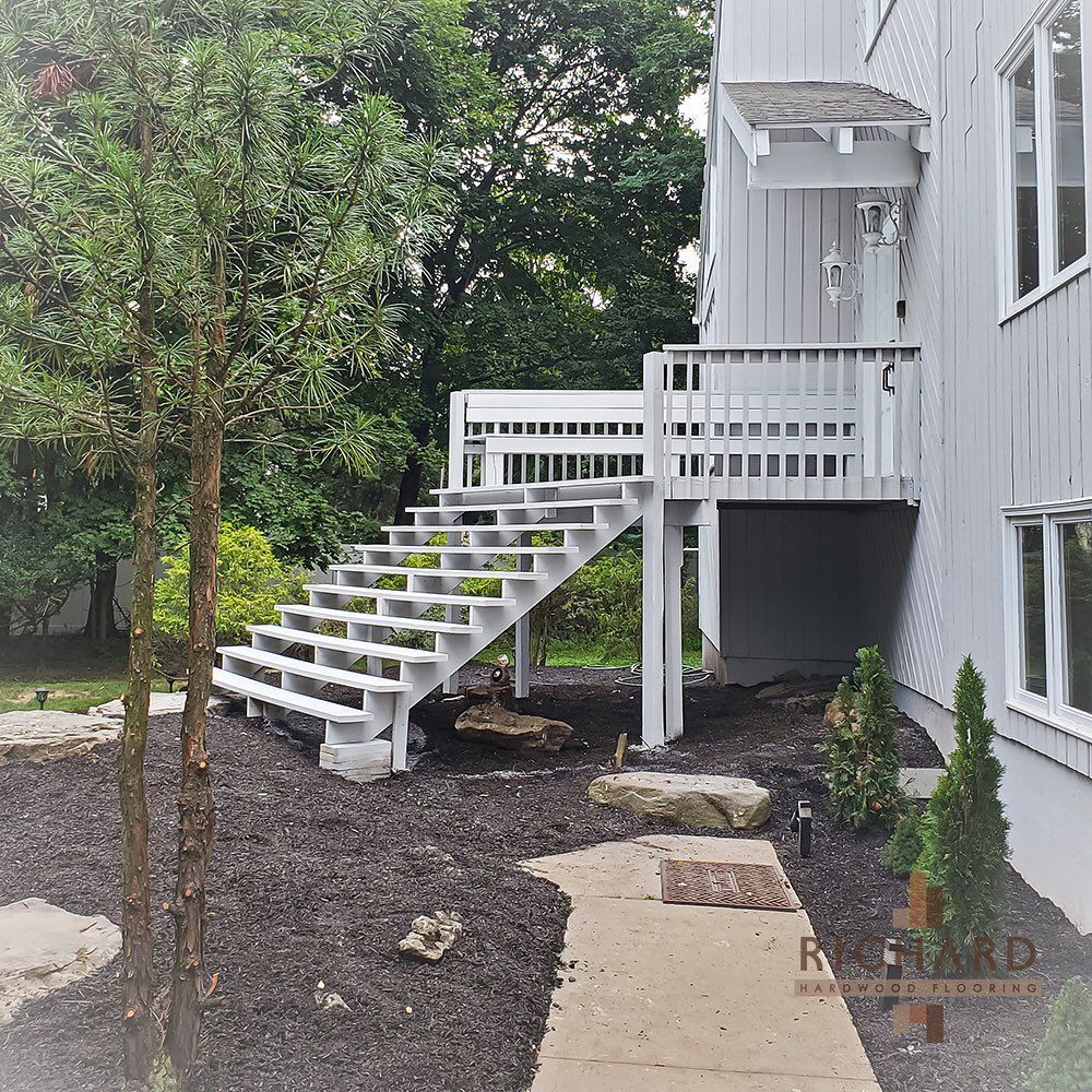 A white outdoor staircase with a wooden railing attached to the side of a grey house, set in a landscaped yard.