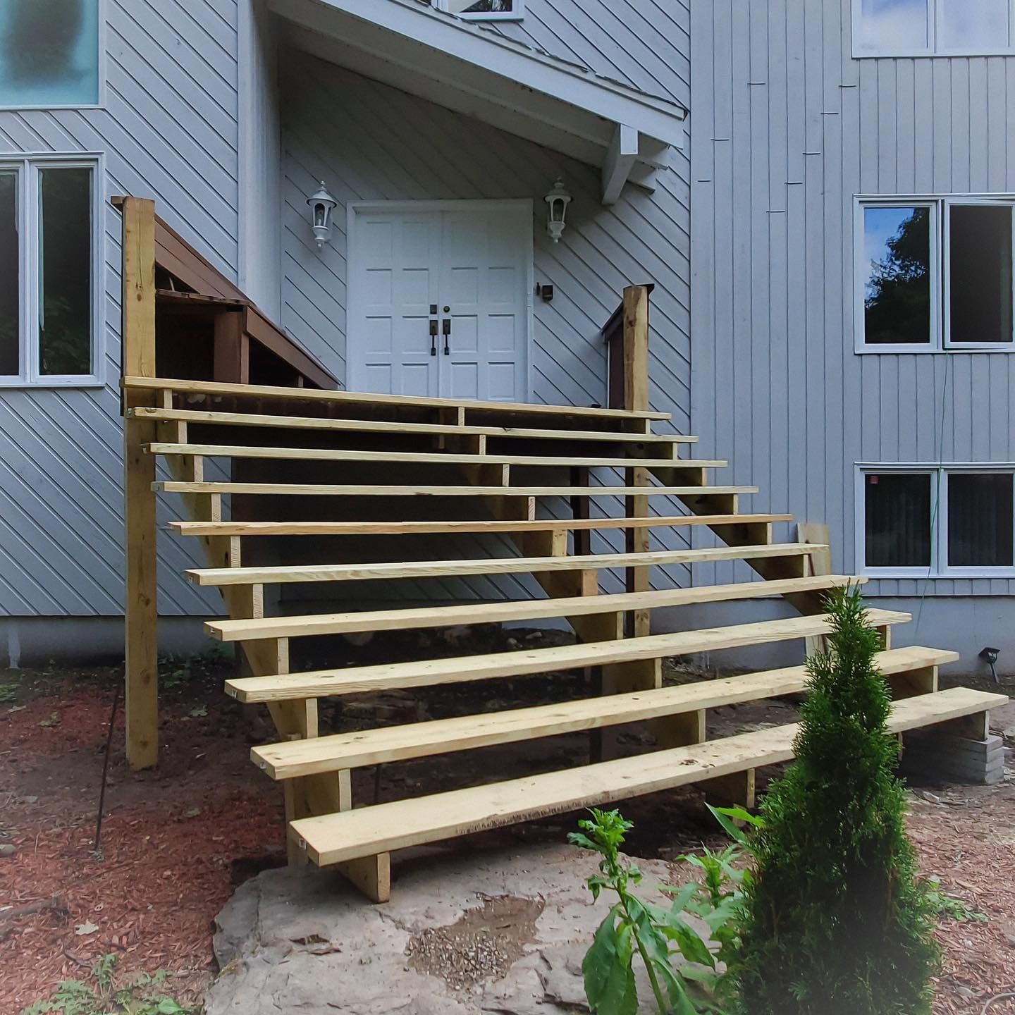 A wide set of unfinished light-wood stairs leading up to a double-door entrance on the front of a light gray house.