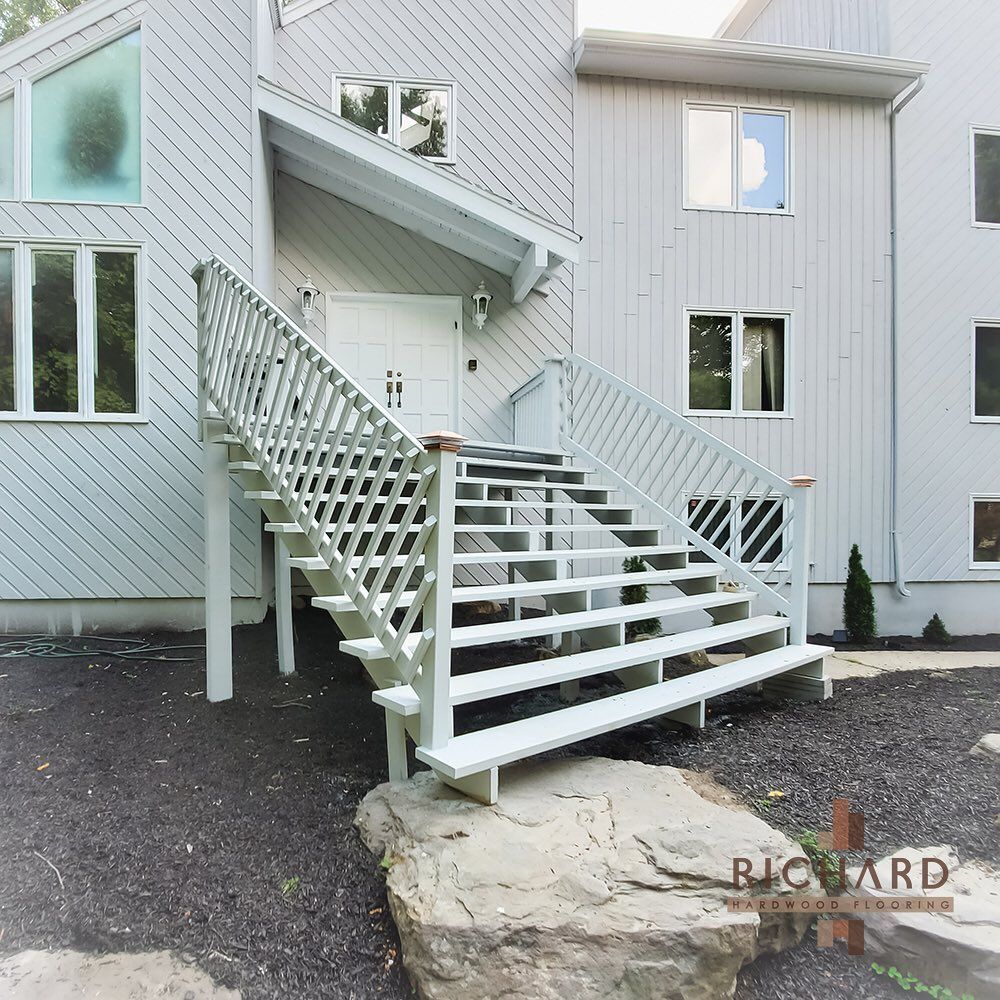 White wooden stairs with lattice railings lead to the entrance of a light-gray house with vertical siding and large windows.