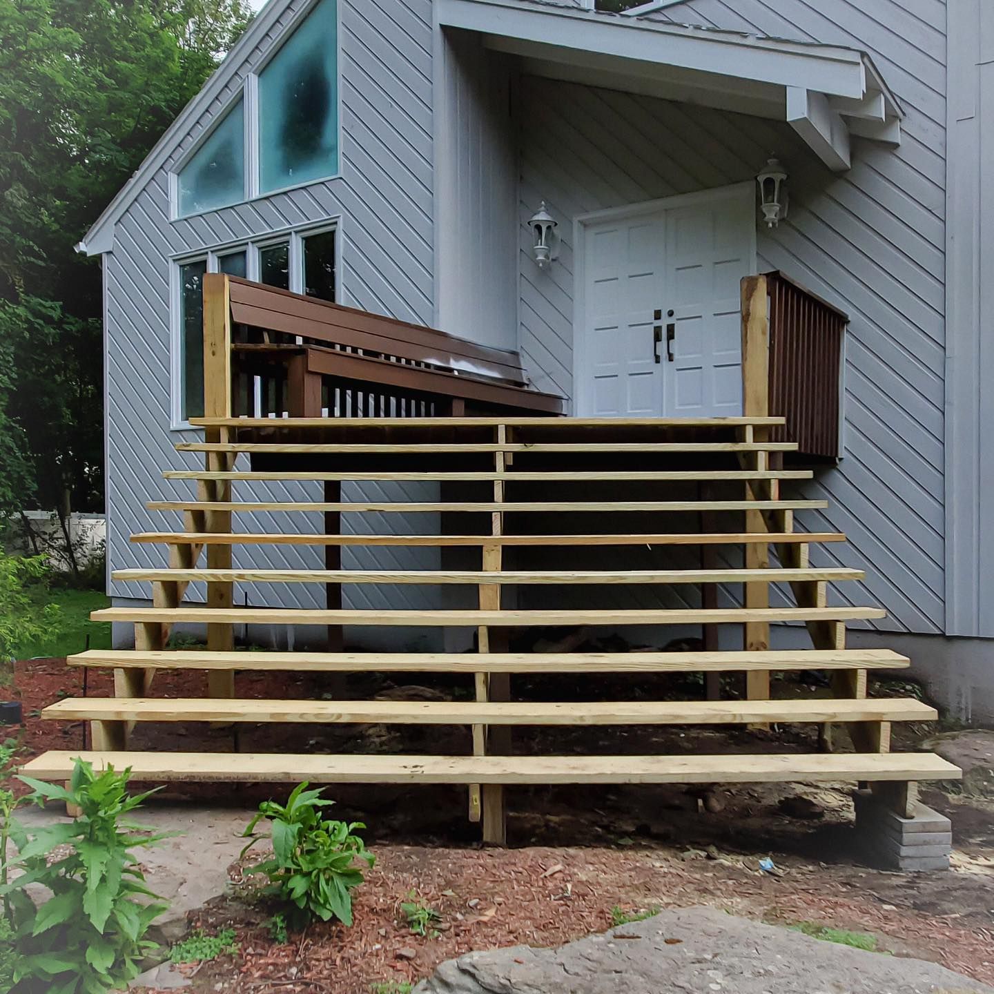A set of newly constructed wooden stairs leading to the double front doors of a light grey, wood-sided house.