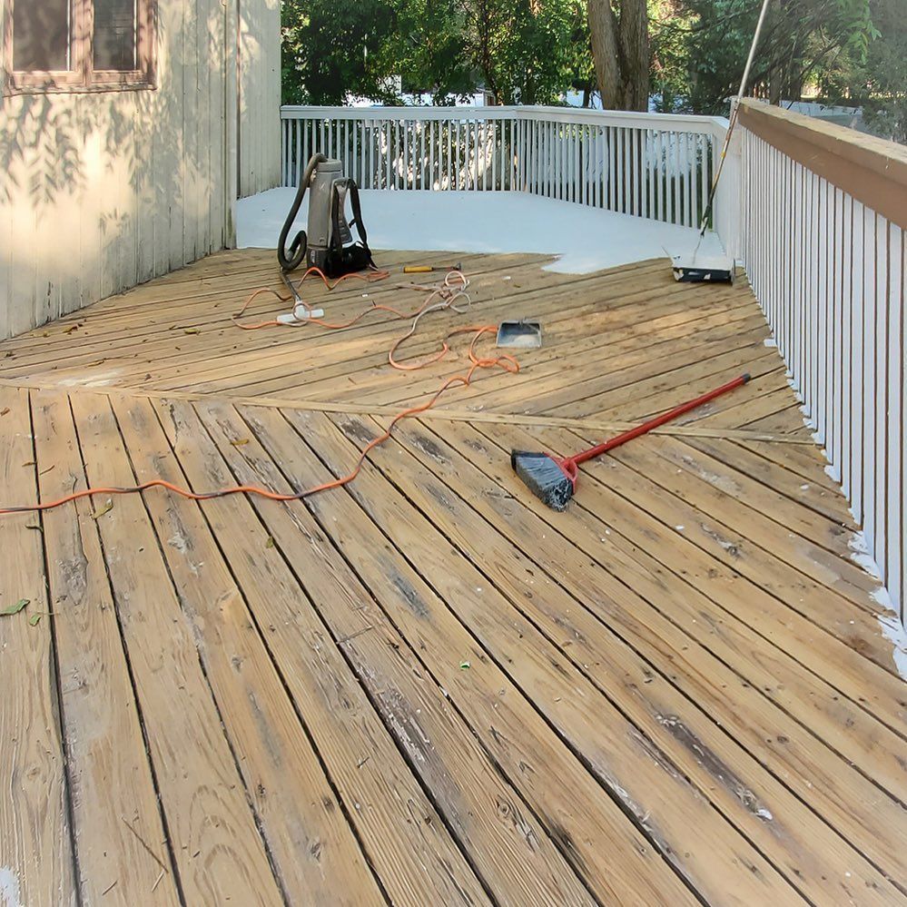 A partially painted wooden deck with a paint sprayer, broom, and orange extension cord scattered across the boards.