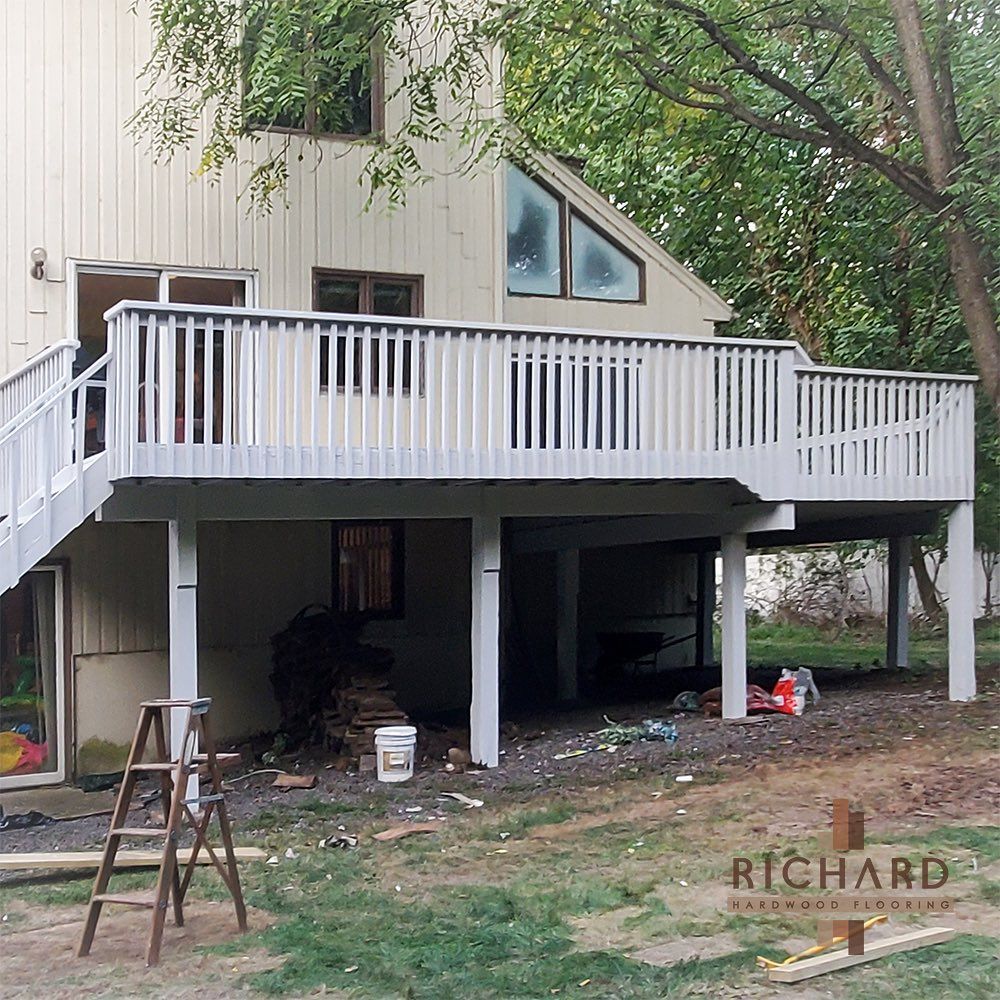 A beige two-story house with a large, elevated light gray wooden deck supported by posts, with a ladder in the yard.