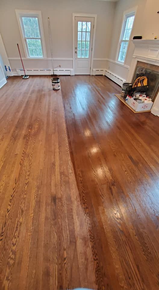 A room showing a hardwood floor mid-refinishing, with one side sanded and light, and the other side stained and glossy.