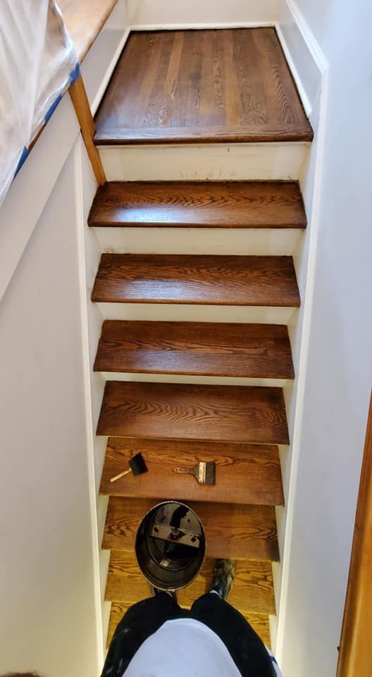 A person standing at the bottom of a staircase with stained wooden steps, a paint can, and brushes.