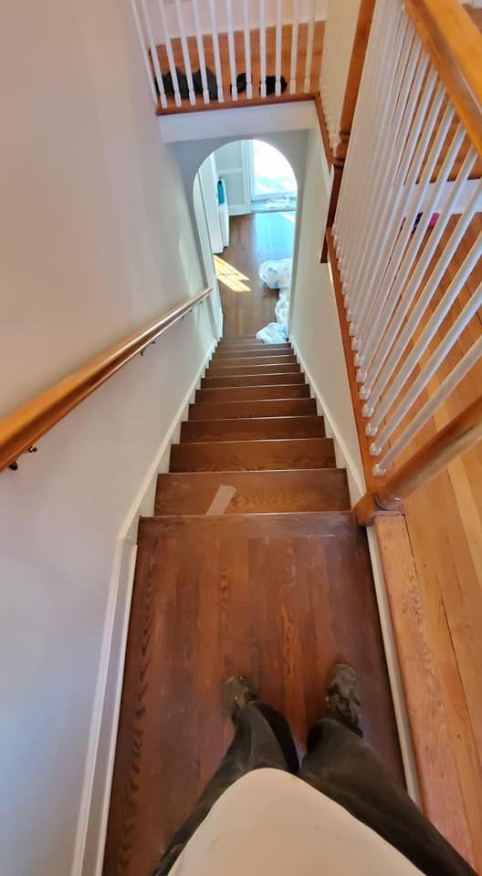A first-person view looking down a narrow wooden staircase with a handrail on the left and a white railing on the right.