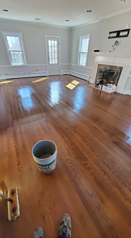 A freshly stained and varnished wooden floor in a living room with white walls, a fireplace, and a bucket of supplies.