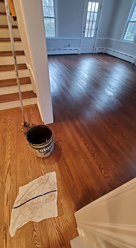 A stained wooden floor in a house transition, featuring a paint bucket and a cloth, with half the floor darker than the rest.