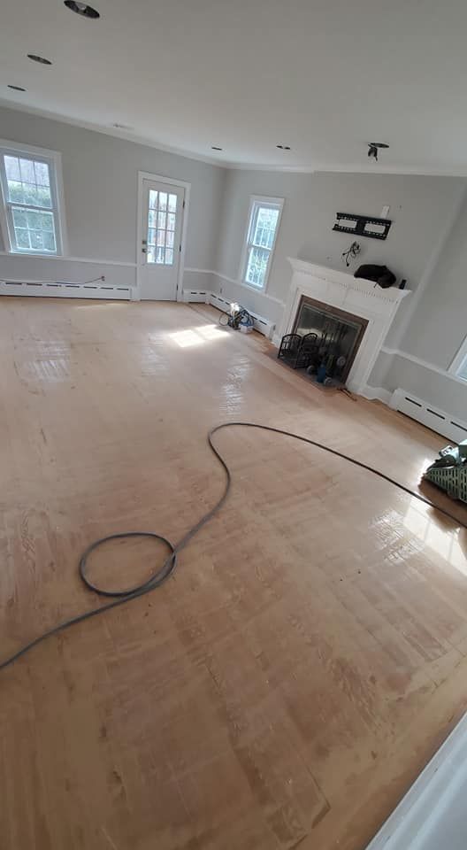A room with newly sanded, unfinished hardwood floors, a white fireplace, neutral grey walls, and a door to the outside.