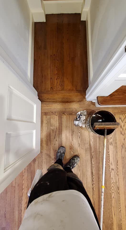 A person stands on a wooden floor in a hallway next to a paint bucket and roller, with a darker wood transition ahead.