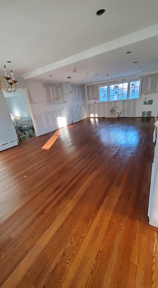An open-plan room with polished hardwood floors, white walls in progress, and natural light streaming from large windows.