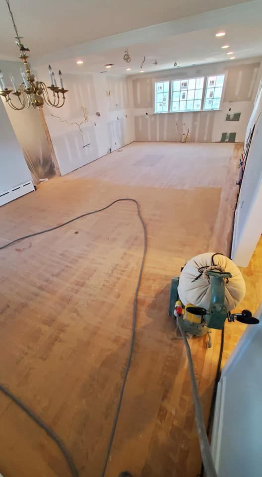 A floor sander sits on an unfinished wood floor in a room undergoing renovations with bare walls and a chandelier.