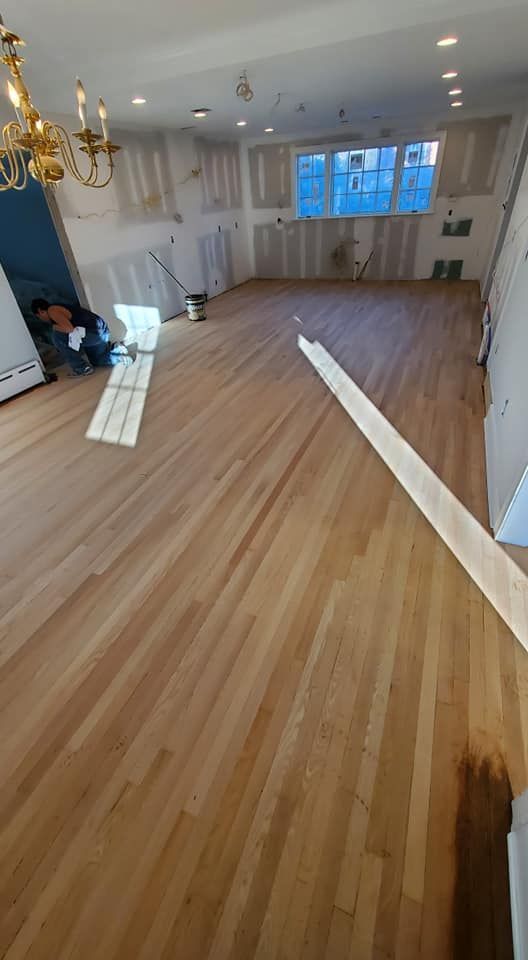 A worker installs hardwood flooring in a room with light wood floors, an ornate chandelier, and unpainted wall panels.