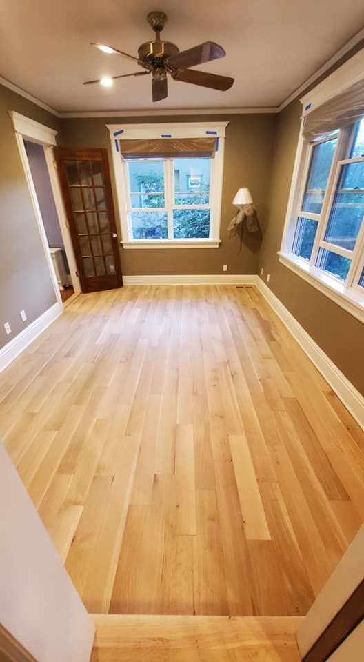 A vacant room with light hardwood floors, taupe walls, a ceiling fan, and two windows.