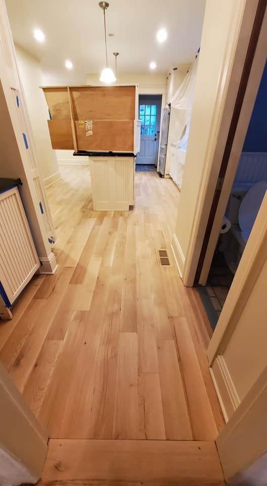 A view through a doorway into a home kitchen with light wood floors, a central kitchen island, and a distant doorway.