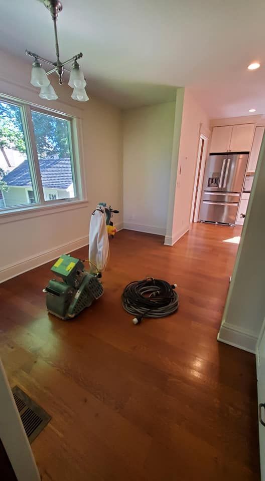 A hardwood floor being refinished with a drum sander and a coiled cord in an empty dining room near a kitchen.