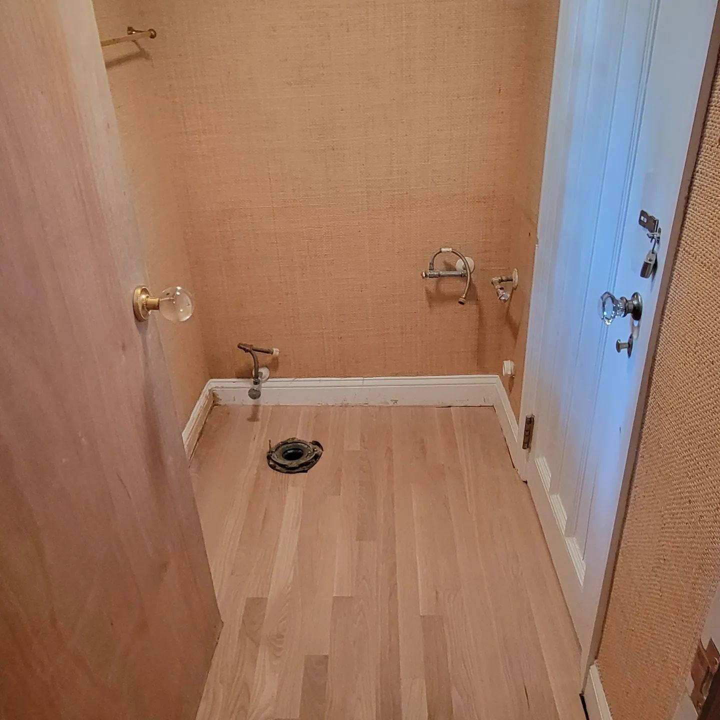 A small, unfinished bathroom with light wood flooring, a toilet flange in the floor, and exposed plumbing on the wall.