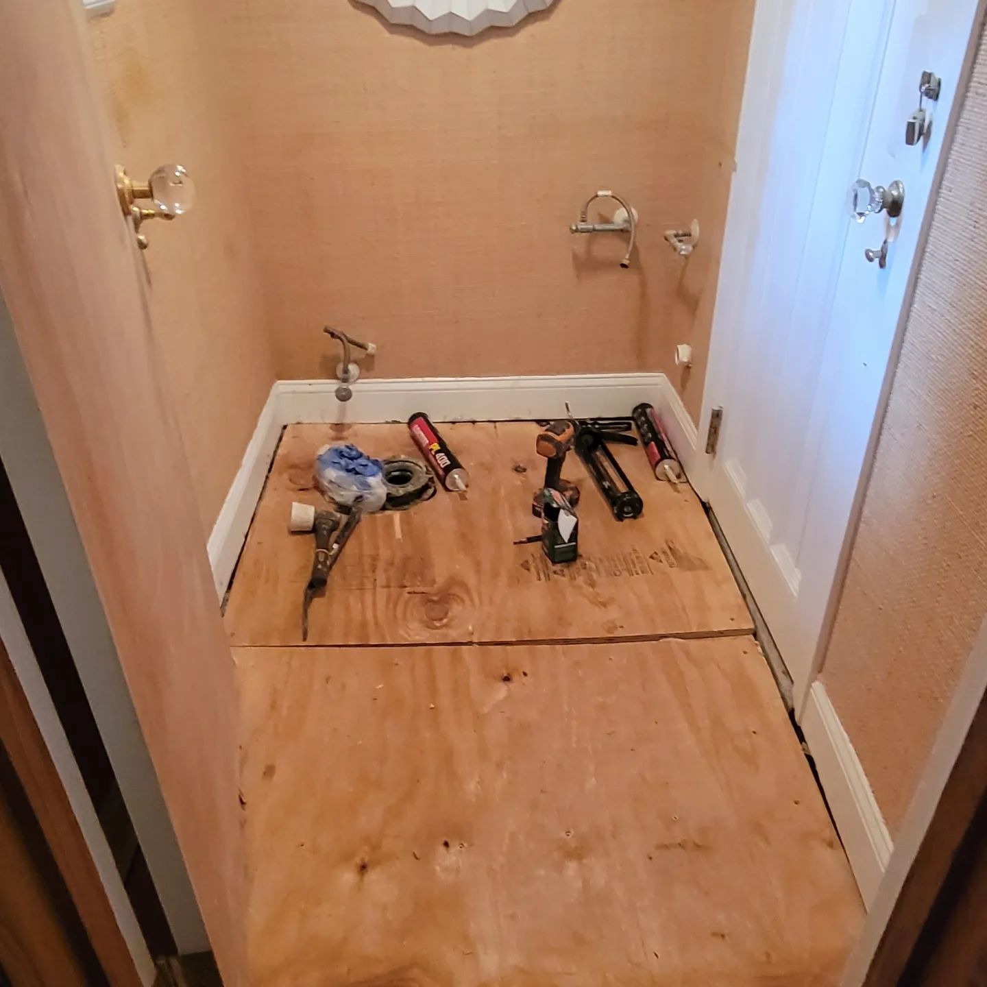 A partially renovated bathroom with plywood flooring, tools, and construction materials scattered on the floor.