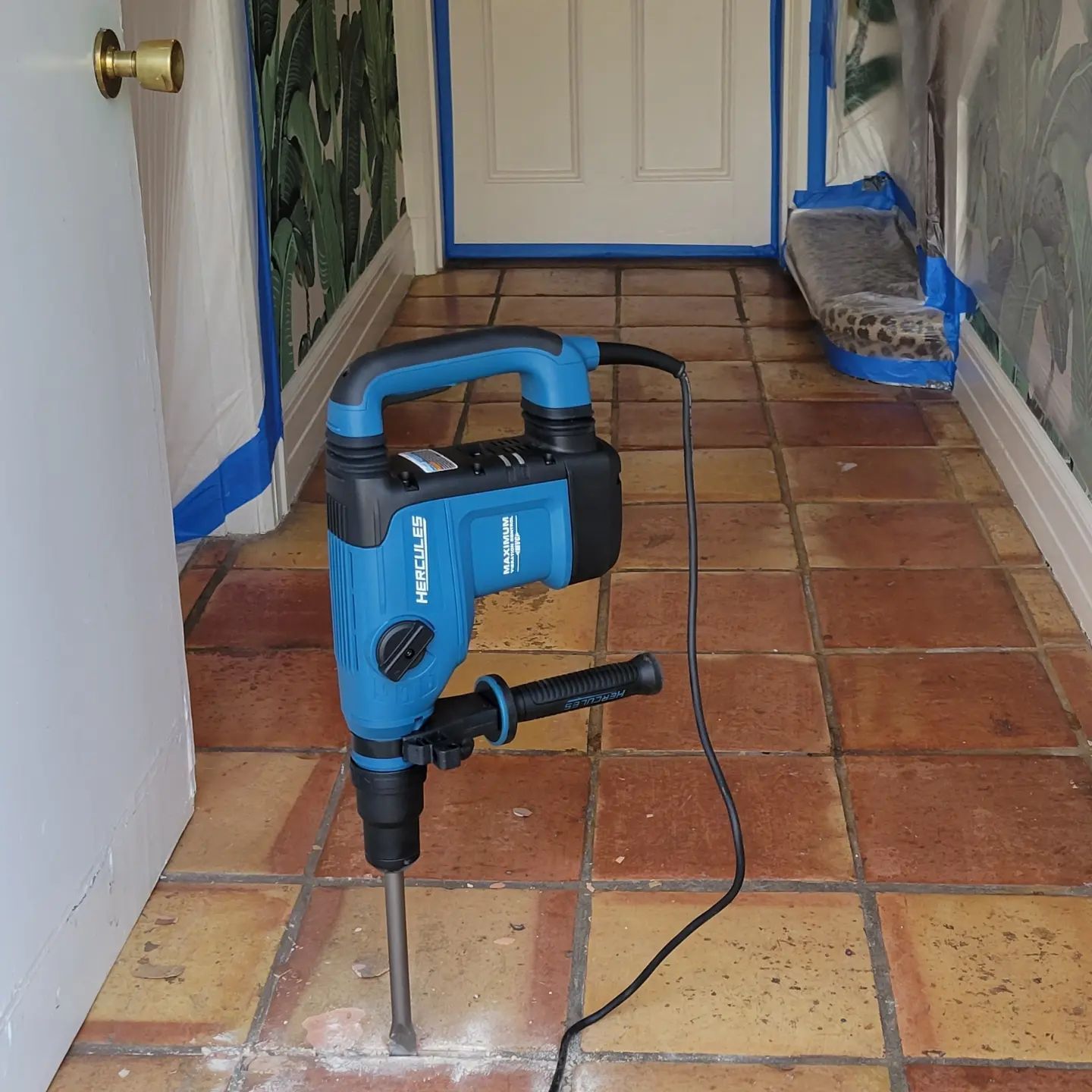 A blue Hercules demolition hammer stands upright on a tiled floor in a hallway during a home renovation.